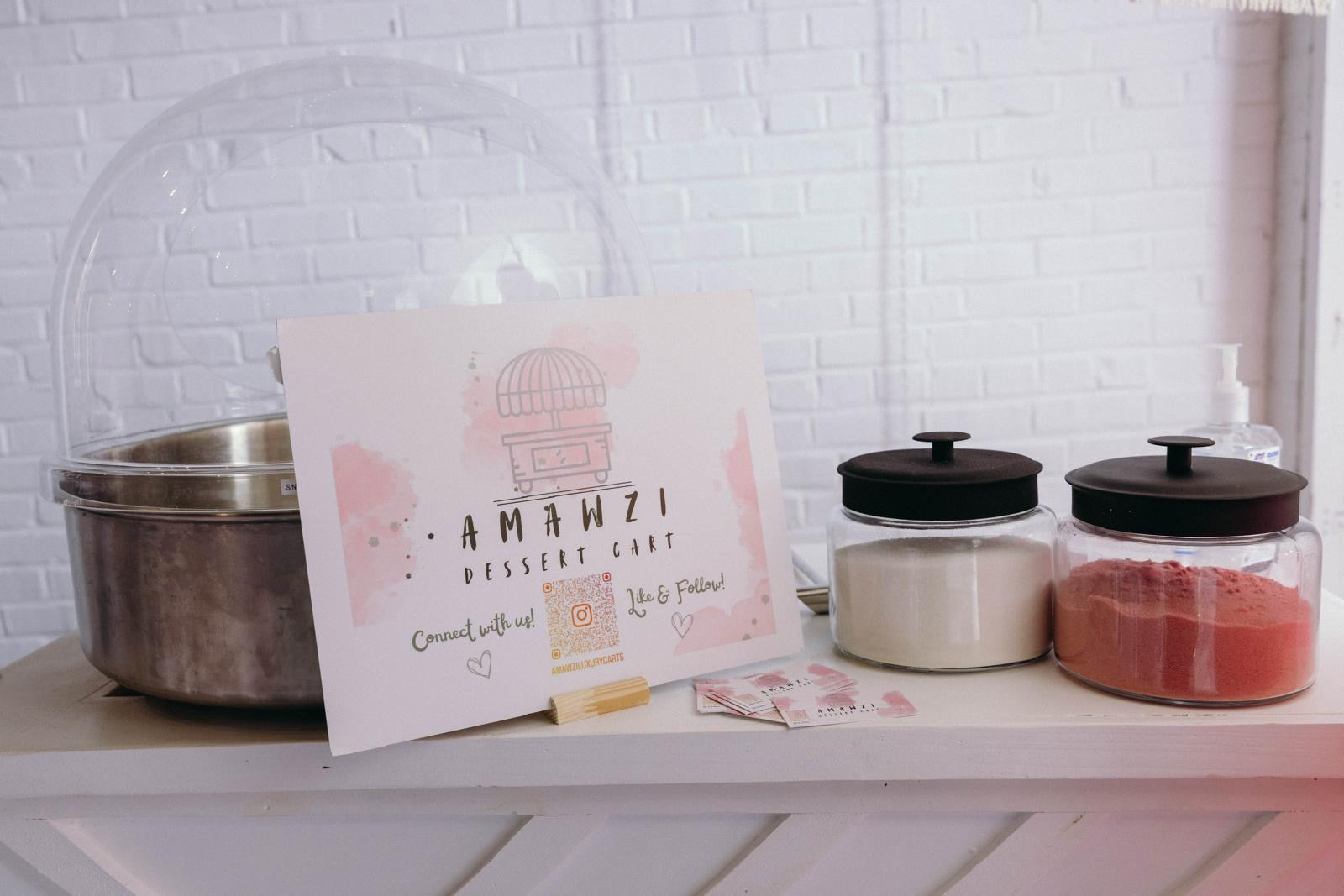 Amanzi Dessert Cart sign on a shelf with jars and a metal pot against a white brick wall.