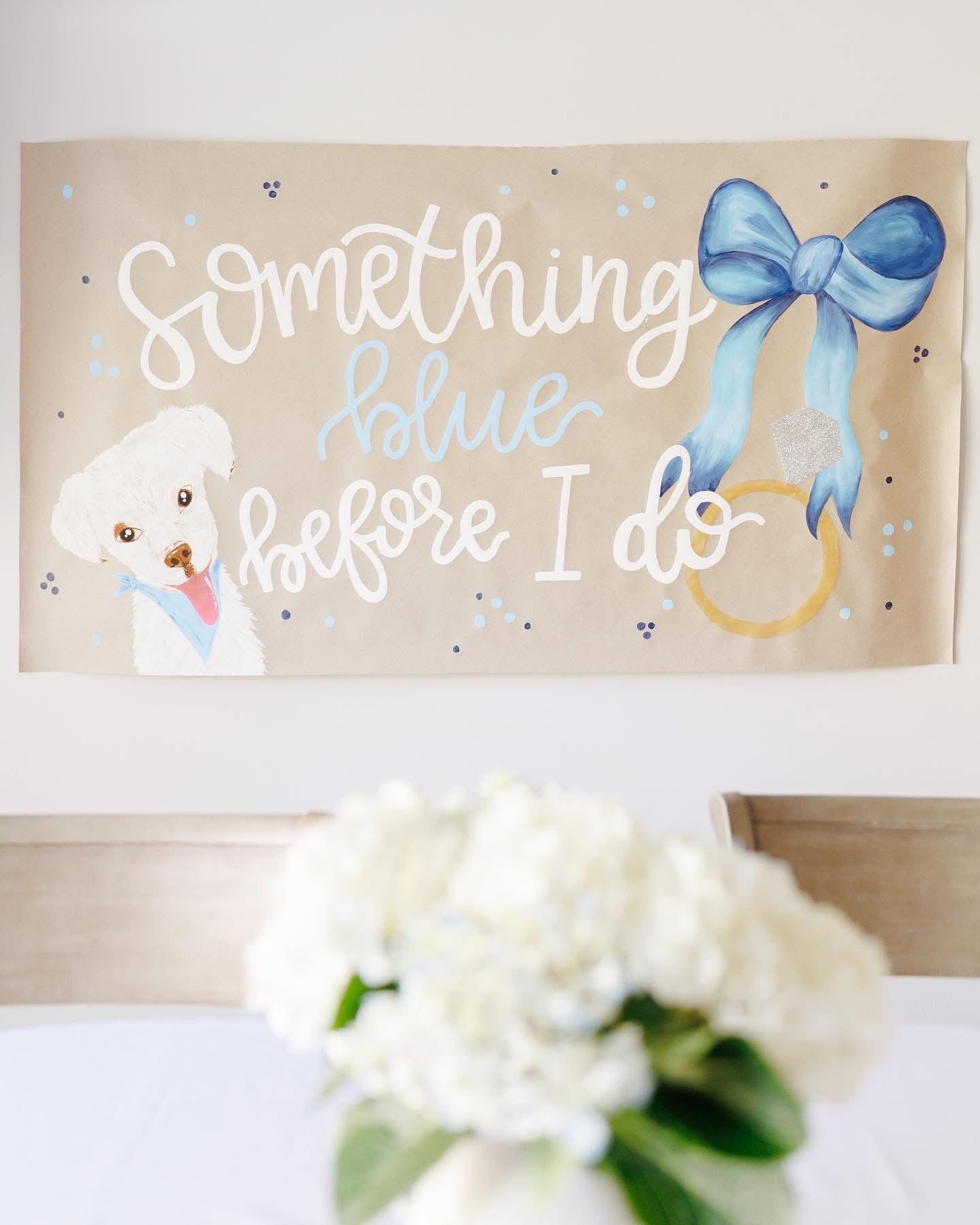 Decorative sign with a dog, blue bow, and ring, with 'Something Blue Before I Do' text, placed above a bouquet of flowers.