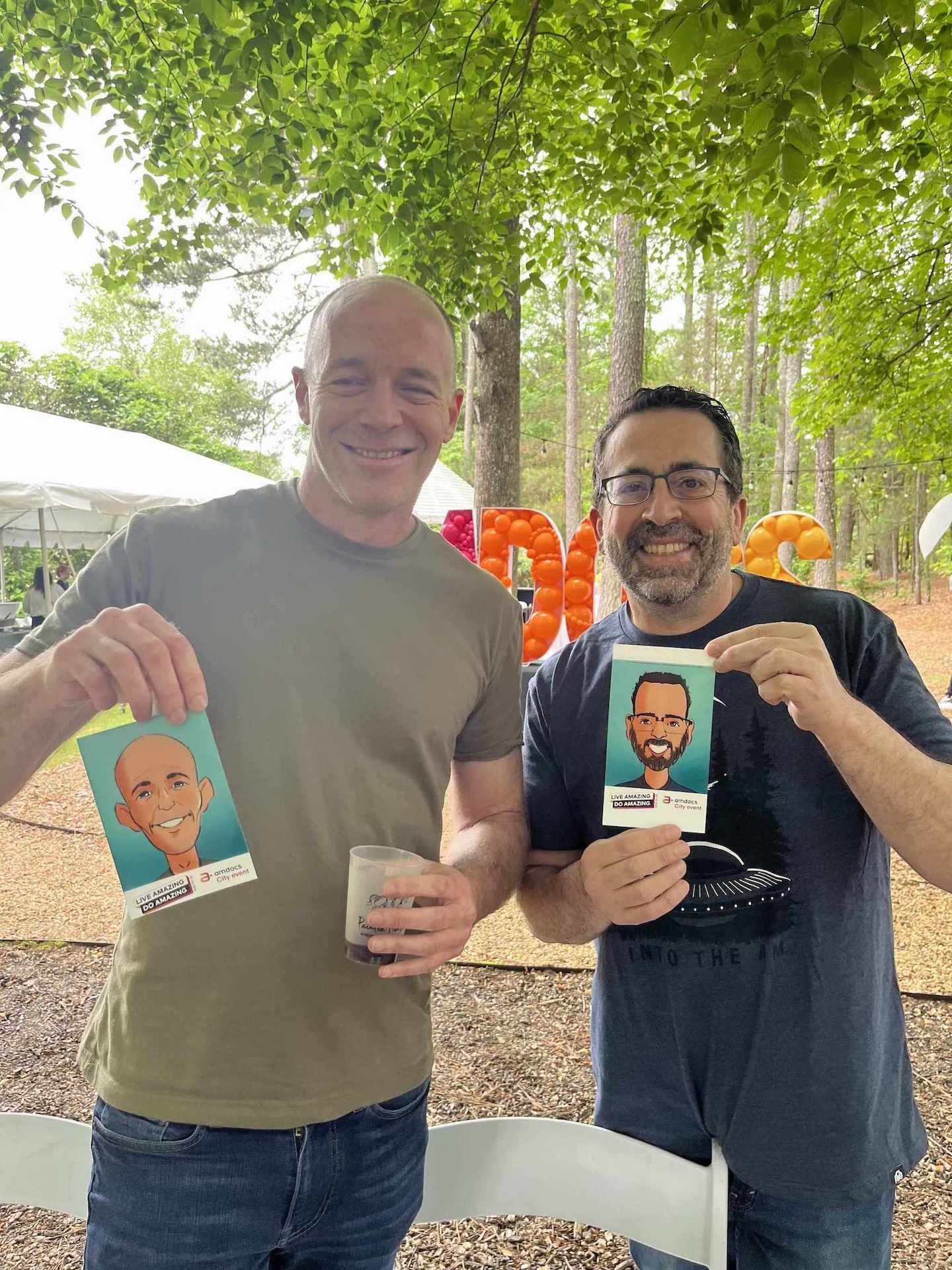 Two men holding up cards with digital caricatures in Atlanta of themselves outdoors under a tree.