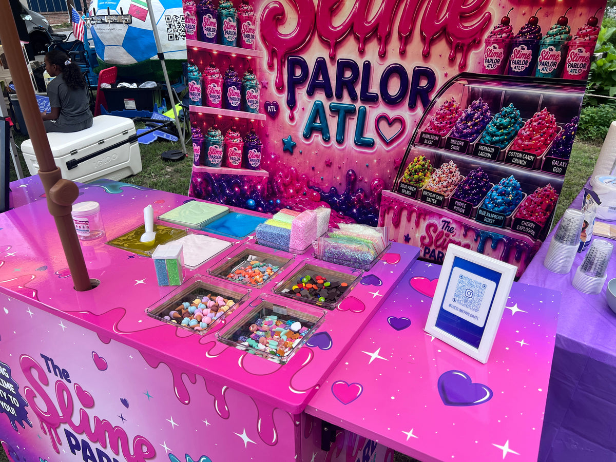 A pink slime party in Atlanta at an outdoor event, featuring a decorated counter with bins of colorful slime, candy, and supplies, a large backdrop reading 'Slime Parlor ATL,' and display cases filled with colorful slime samples.