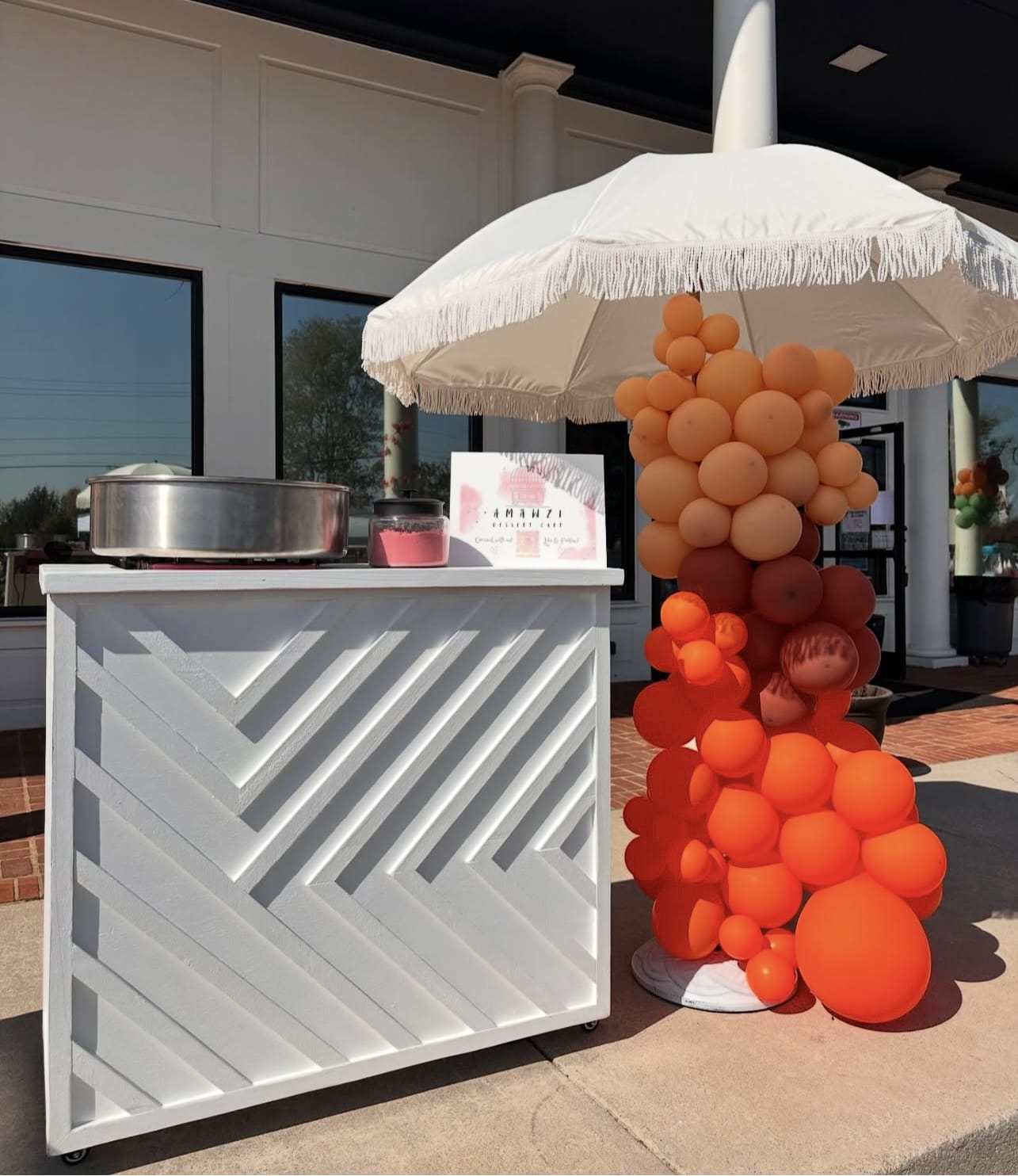 White outdoor dessert bar cart with geometric design, orange balloon sculpture, and white umbrella.