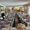 Dining room with tables set for a formal event, featuring floral centerpieces and purple table runners.