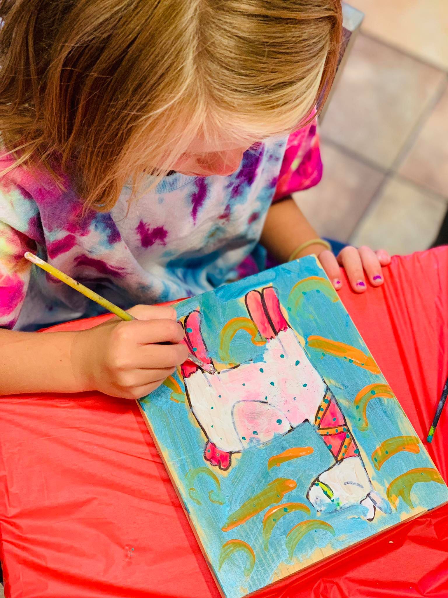 Child painting a colorful picture of a cartoon character on a red blanket at a painting birthday party.