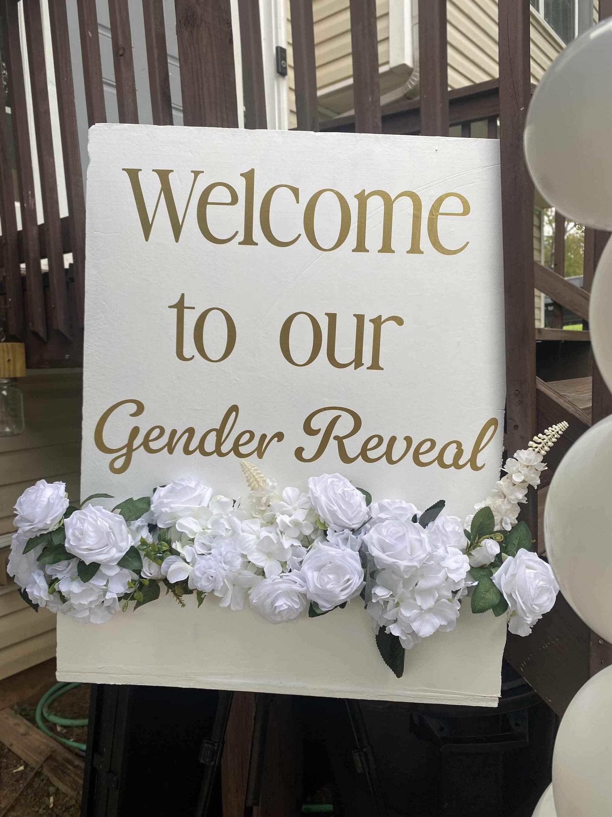 Gender reveal sign with white flowers and balloons on a wooden deck.