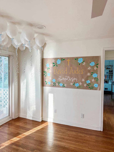Painted banner with floral designs and text on a wall in a room with wooden flooring and balloons.