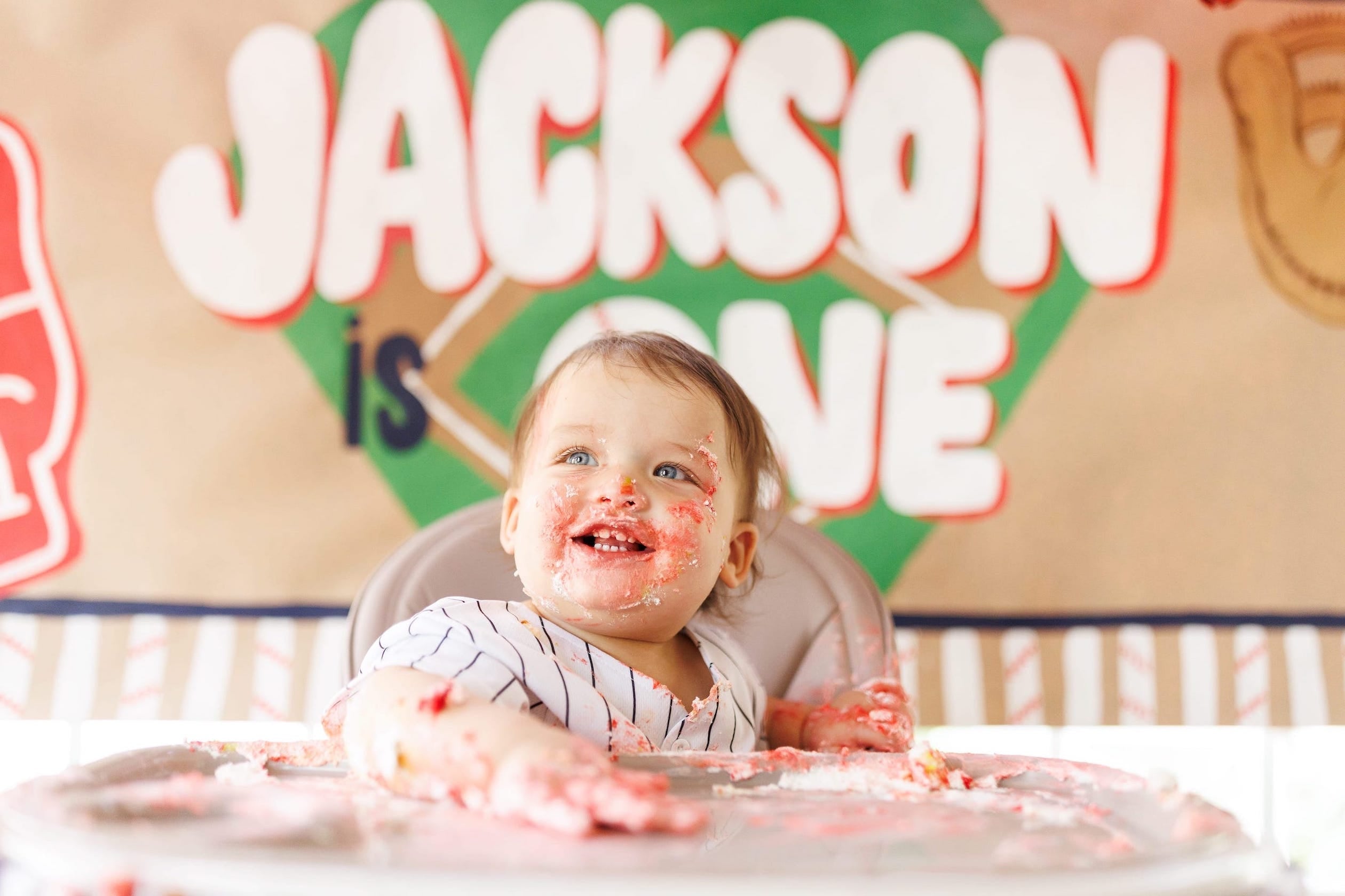 Baby with cake on face in front of a colorful custom painted banner with 'Jackson is one' 