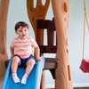 Child sitting on a slide in a playground setting with a swing in the background.