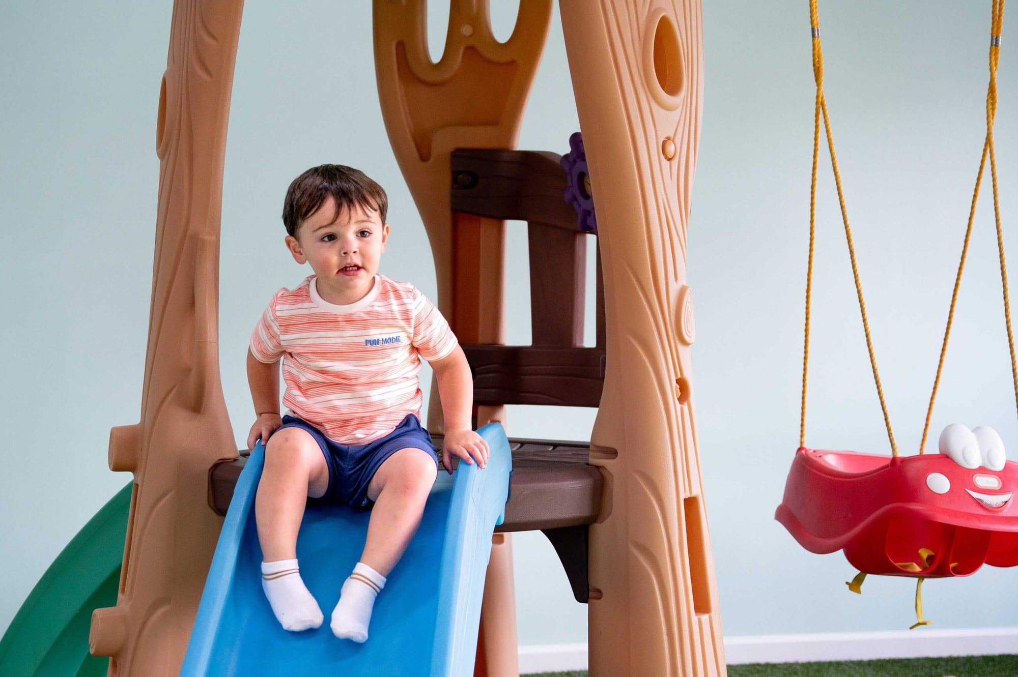 Child sitting on a slide in a playground setting with a swing in the background.
