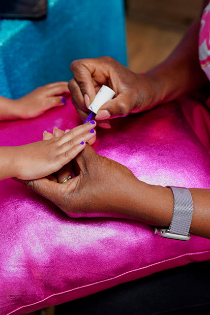 Girls getting her nails painted with purple nail polish on a pink surface.