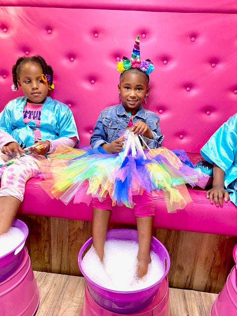 Children in colorful outfits sitting on a pink bench getting pedicures at a spa bus party in Atlanta.