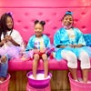 Three children sitting on a pink couch in a spa party bus with soapy feet in buckets, wearing colorful outfits.
