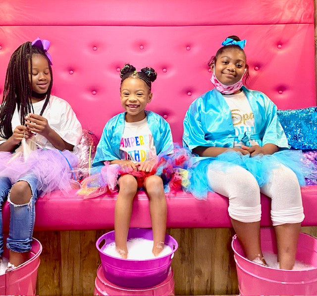 Three children sitting on a pink couch in a spa party bus with soapy feet in buckets, wearing colorful outfits.