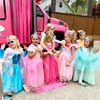 Children in princess costumes standing in front of a pink spa party bus, with a house and trees in the background.