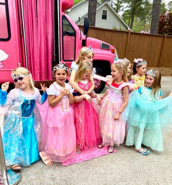 Children in princess costumes standing in front of a pink spa party bus, with a house and trees in the background.