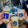 Three bottles and cups of Qamar Coffee Co. held by people outdoors under a blue tent.