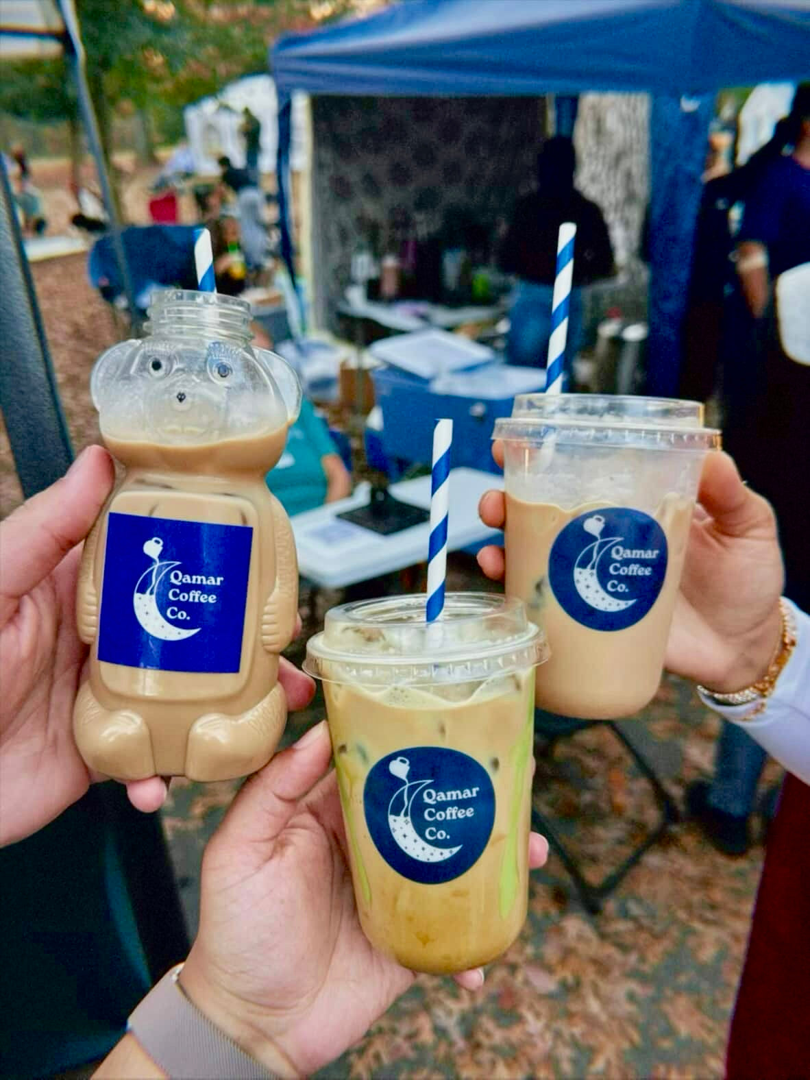 Three bottles and cups of Qamar Coffee Co. held by people outdoors under a blue tent.