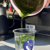 Green macha being poured from a container into a clear cup with a blue label.