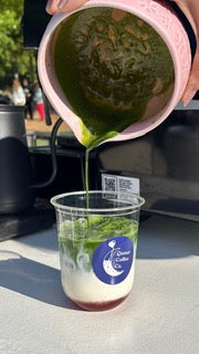 Green macha being poured from a container into a clear cup with a blue label.