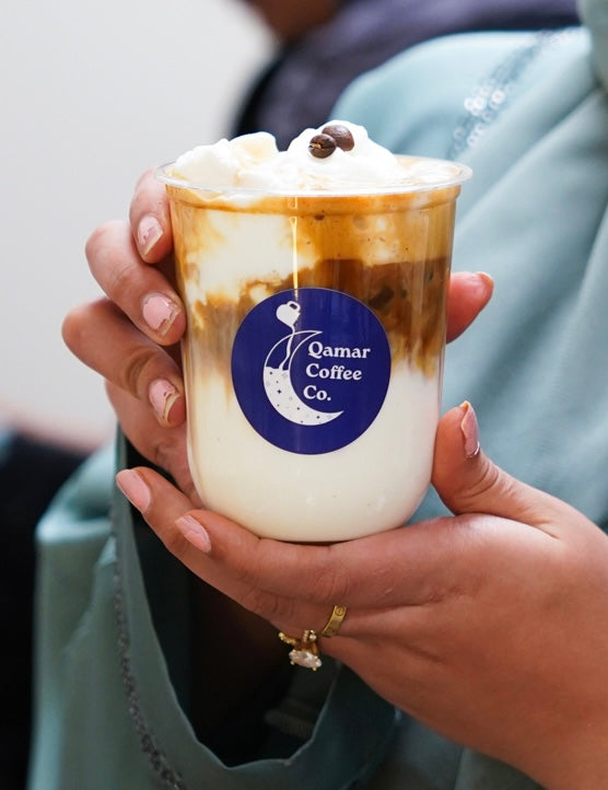 Hand holding a cup of coffee from Qamar Coffee Co. with a visible logo.