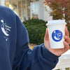 Person holding a white coffee cup with a logo outdoors