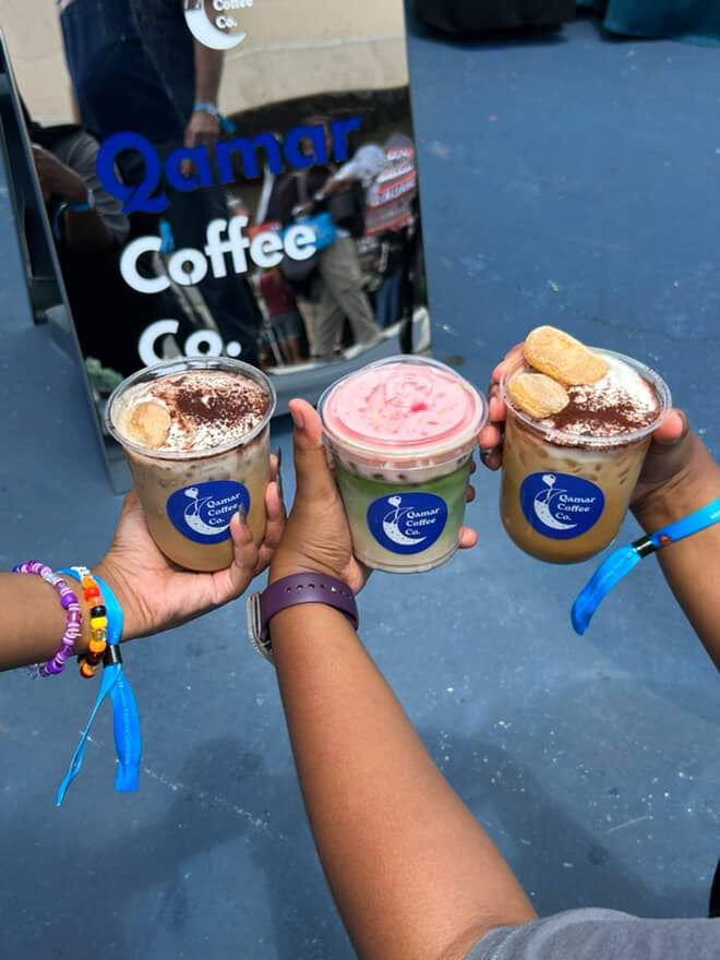 Three people holding coffee drinks from Qamar Coffee Co. with a sign in the background.