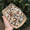 Hand holding a paper container of popcorn with colorful toppings against a green leafy background | Popcorn Cart Atlanta | Confetti Jar