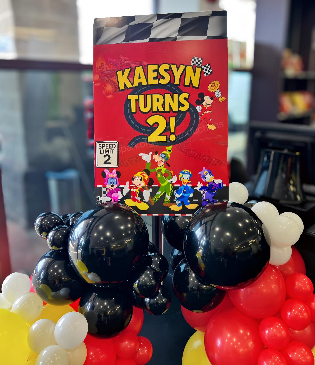 Decorative sign with 'KAESYNturns2!' on a balloon arch