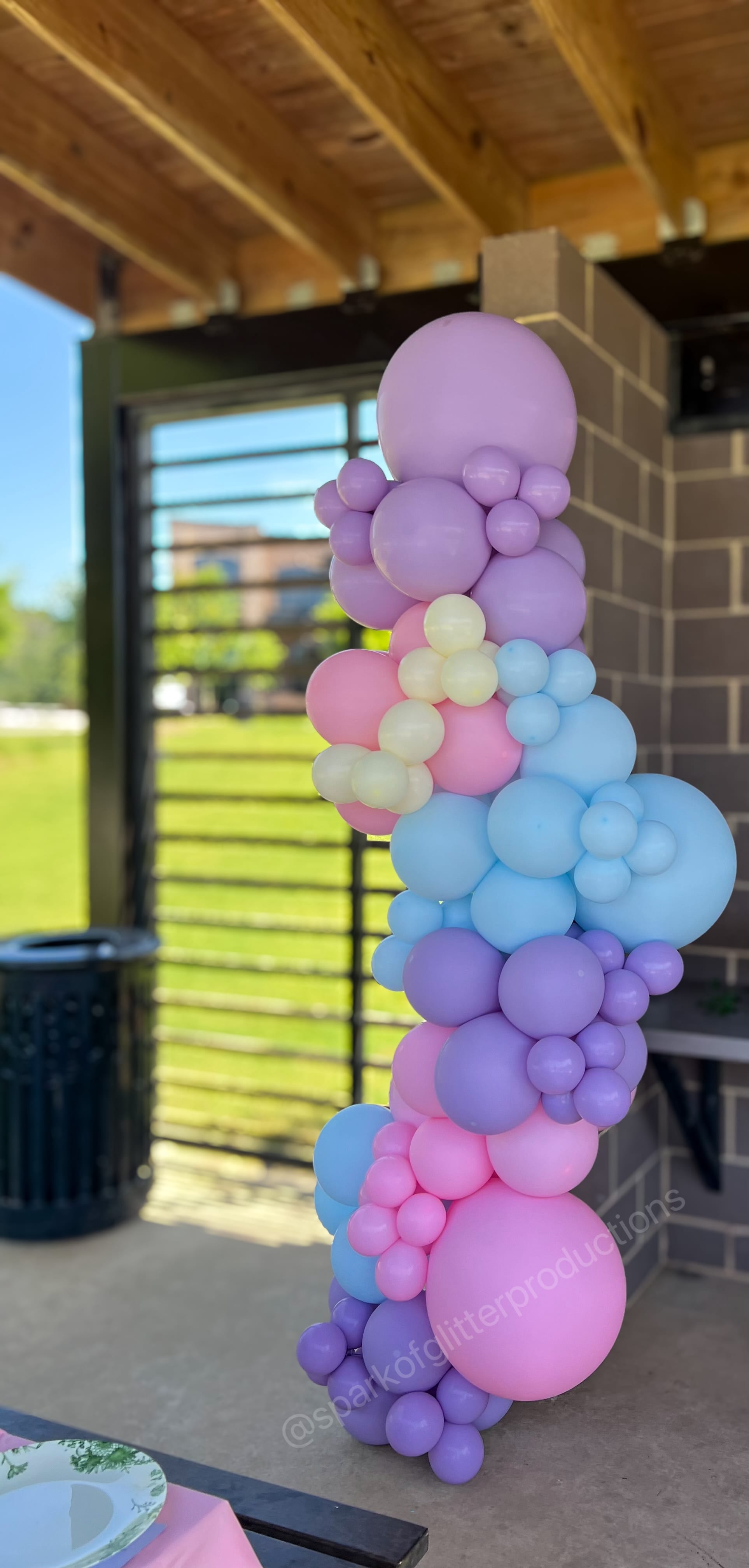 Pastel balloon arch at an outdoor event with a wooden structure and greenery in the background.