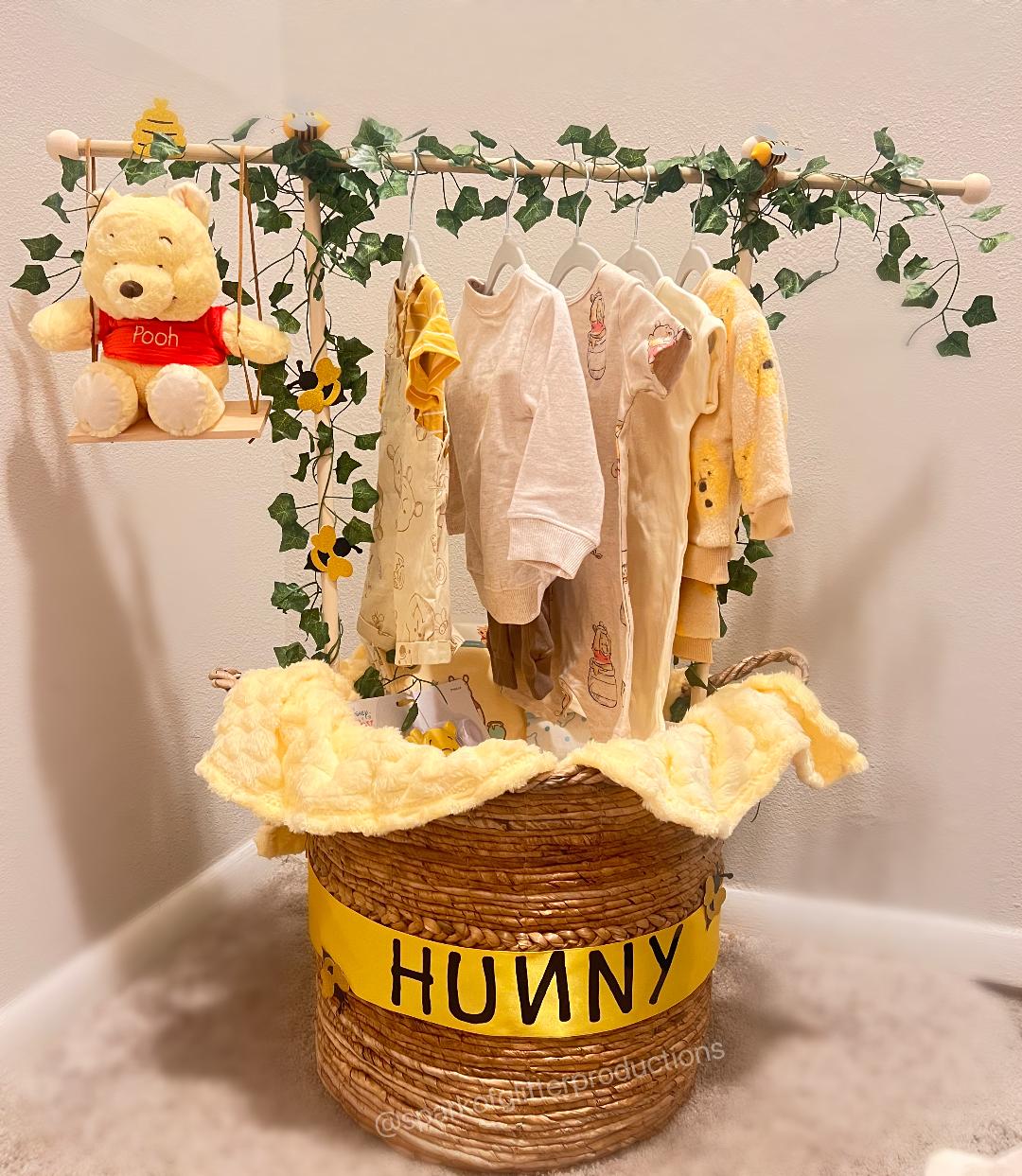 Wicker basket labeled 'Hunny' with baby clothes and a teddy bear on a branch against a white background