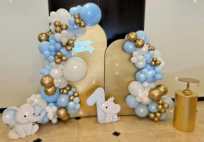 Balloon arch with blue, white, and gold balloons, elephant decorations, and a number '1' on a wooden stand.