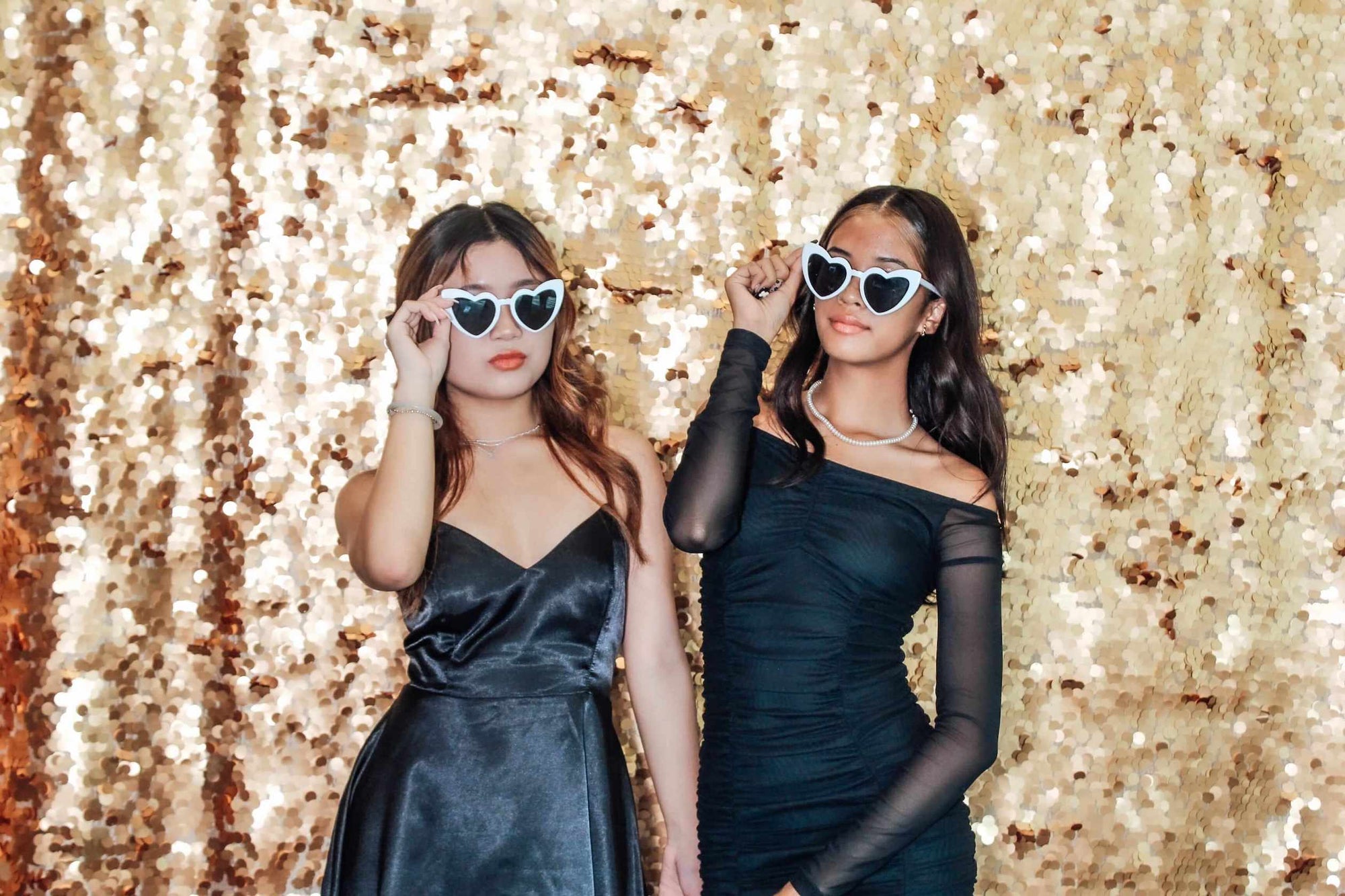 Two women in black dresses with heart-shaped sunglasses against a glittery gold background