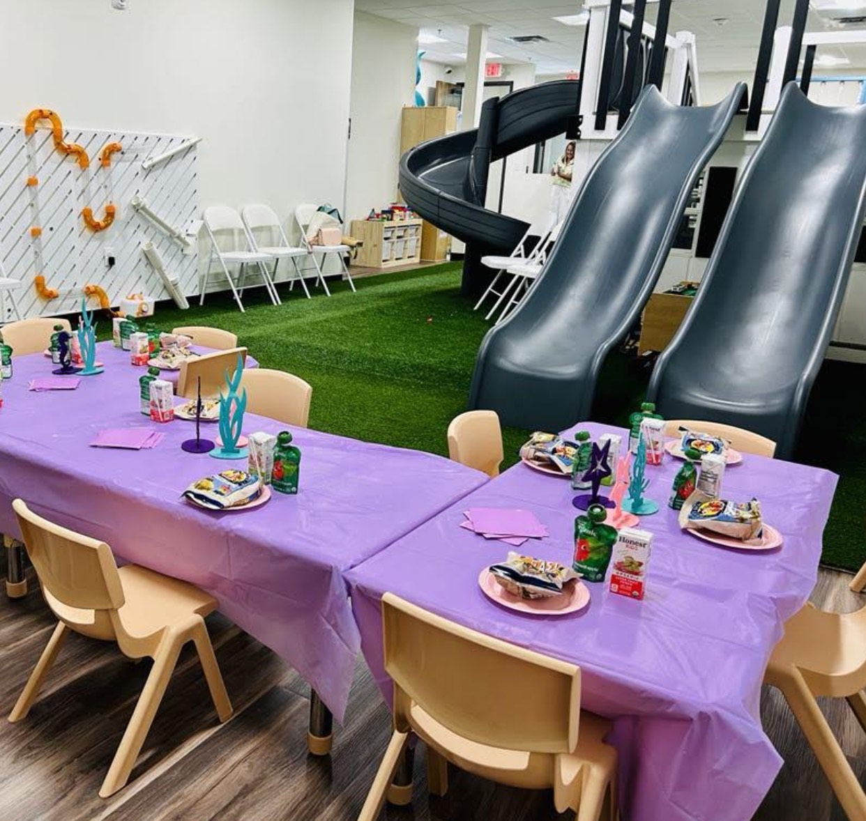 Children's party setup with purple tablecloths, plates, and chairs in a room with slides.