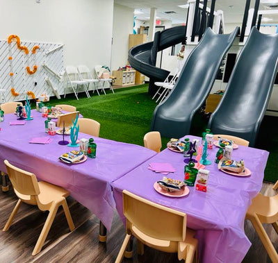 Children's party setup with purple tablecloths, plates, and chairs in a room with slides.