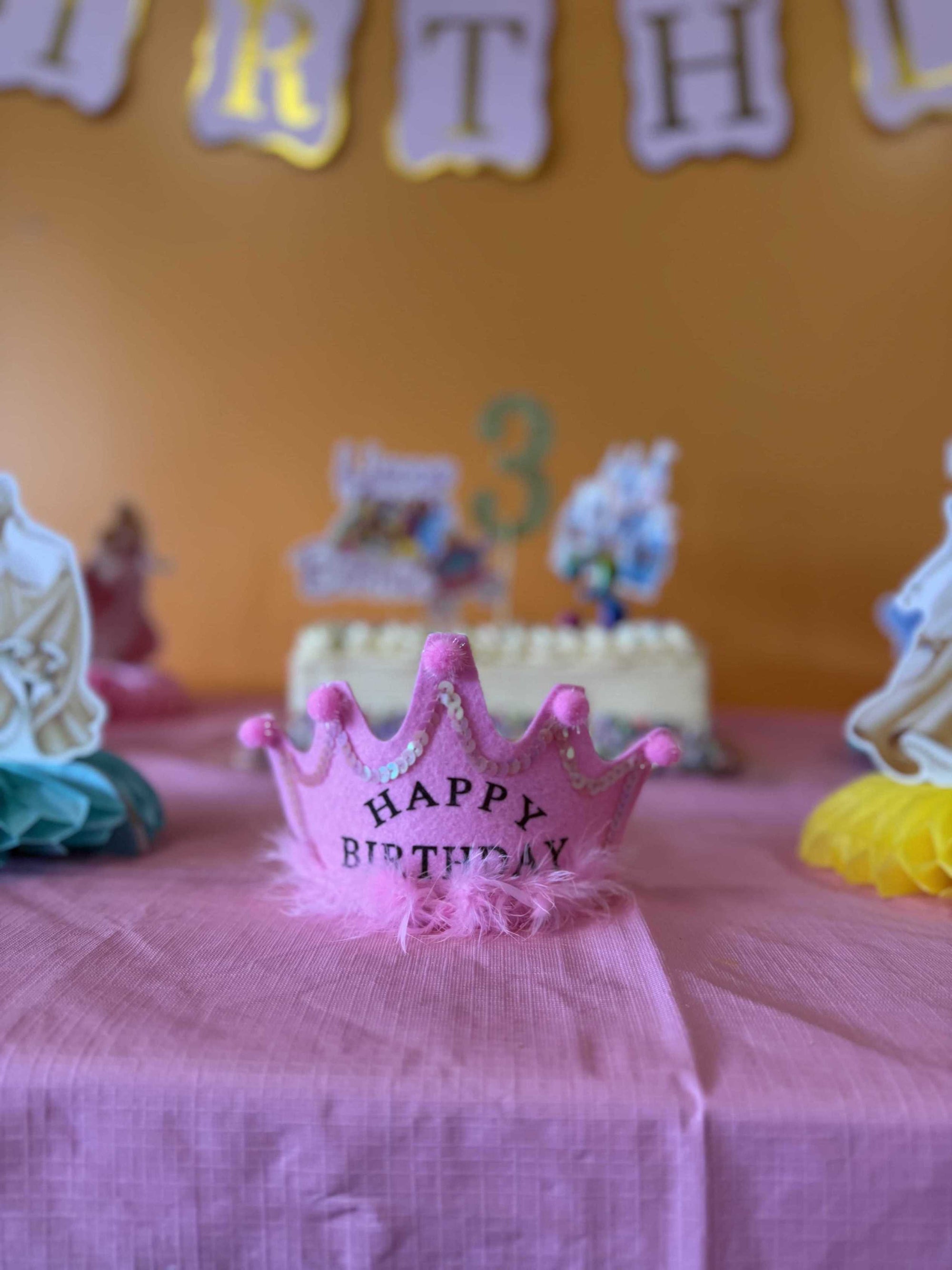 Pink birthday crown on a table with a blurred background of a birthday cake and decorations for a girls birthday party in Johns Creek GA.