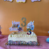 Small birthday cake with number 3 candles on a pink tablecloth against an orange wall with 'Happy Birthday' letters for a toddler birthday party in Johns Creek GA.
