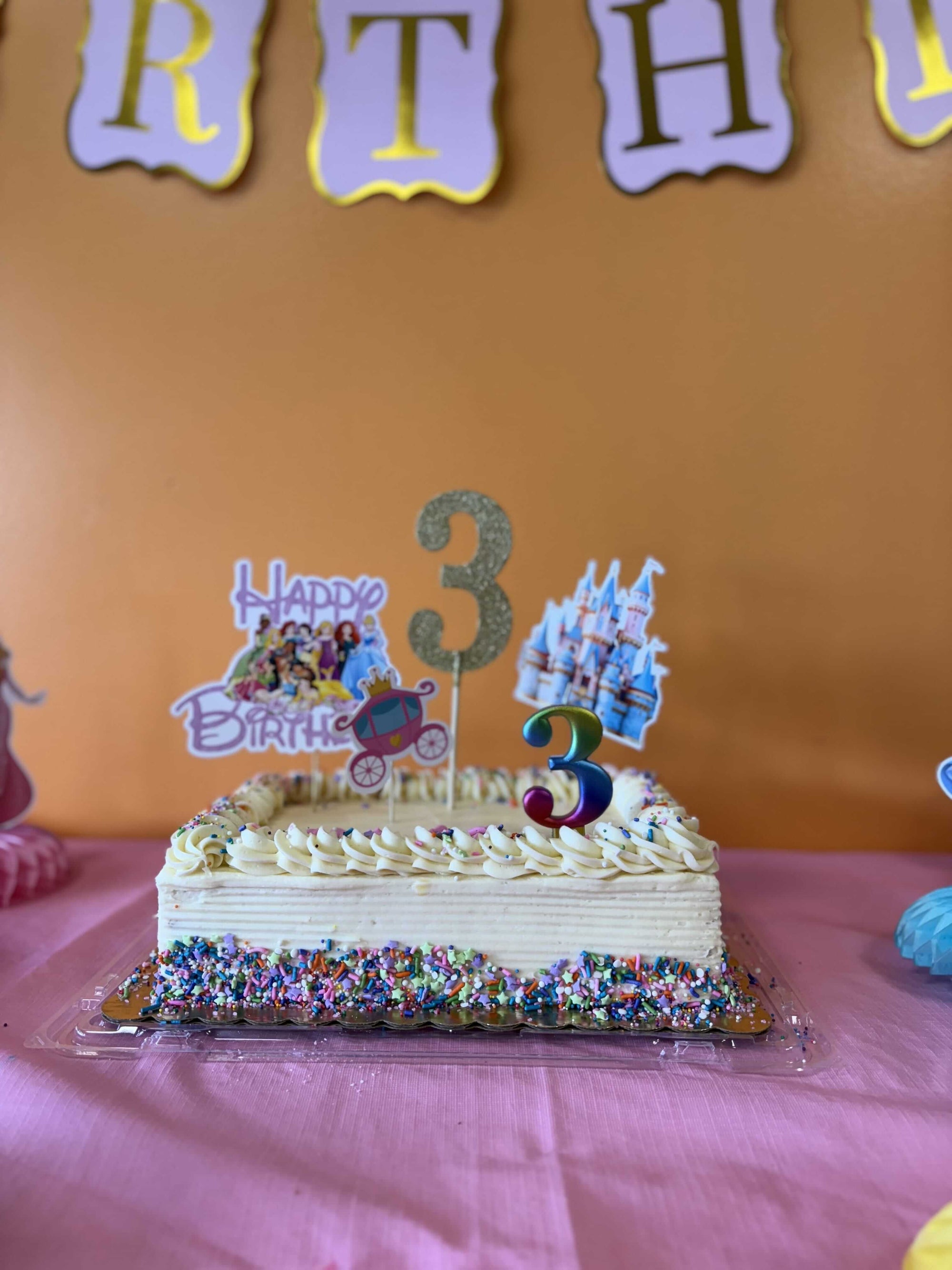 Small birthday cake with number 3 candles on a pink tablecloth against an orange wall with 'Happy Birthday' letters for a toddler birthday party in Johns Creek GA.