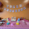 Birthday party setup with cake, decorations, and 'Happy Birthday' banner on a pink tablecloth for a 3 year old birthday party in Alpharetta GA.