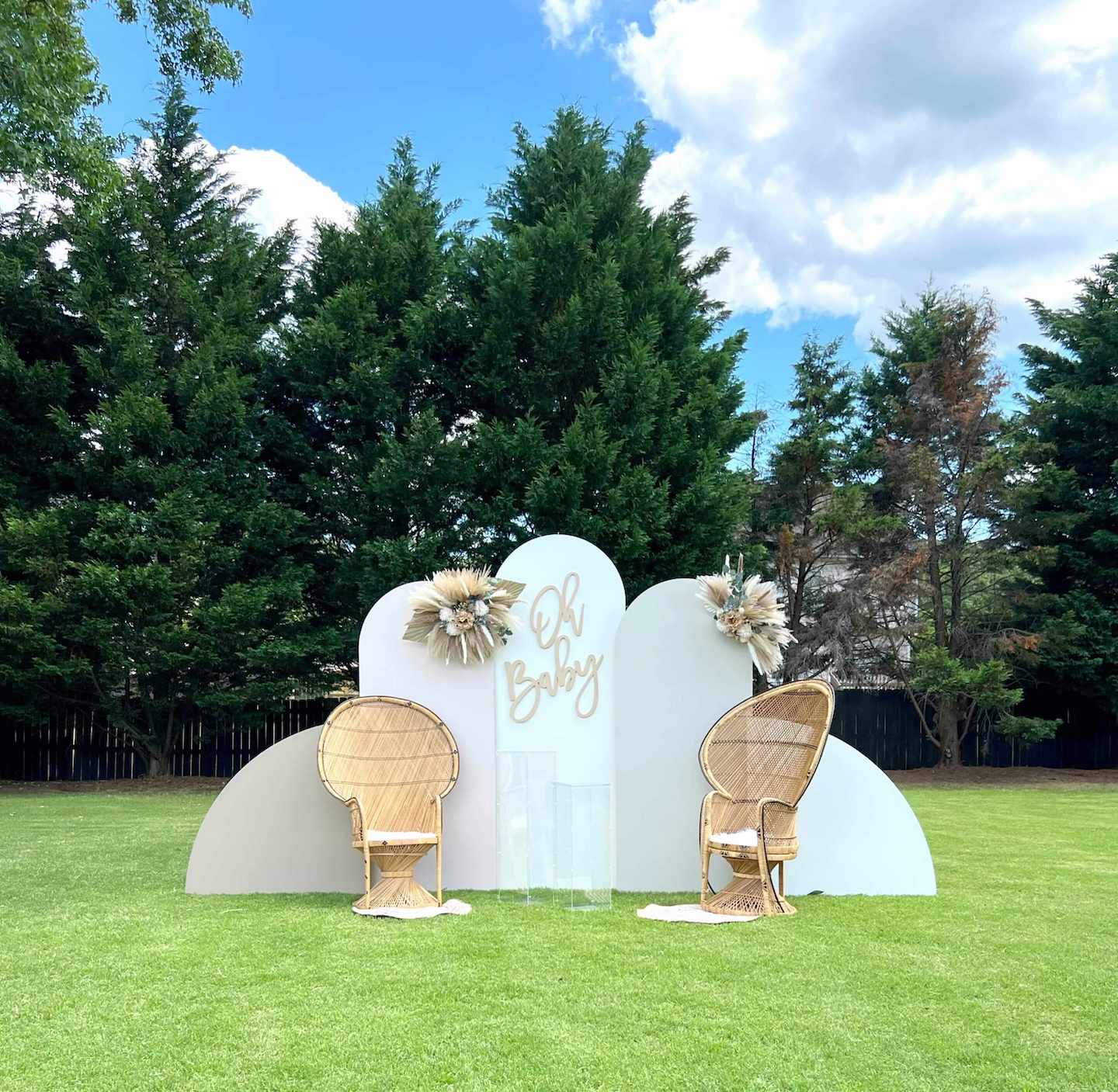 Baby shower backdrop with floral decorations and wicker chairs on a grassy area with trees in the background.