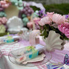 Decorative table setting with flowers and a shell-shaped vase on a pink tablecloth.