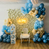 Balloon arch with blue, white, and gold balloons in a room with floral wall and 'Baby' sign.
