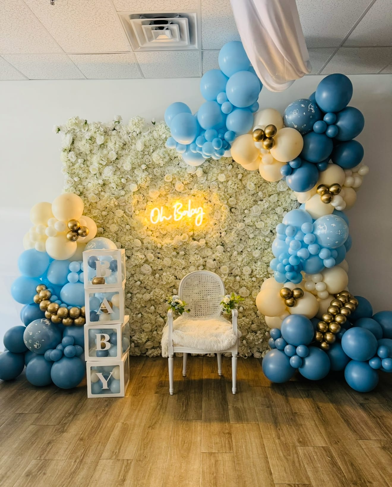 Balloon arch with blue, white, and gold balloons in a room with floral wall and 'Baby' sign.