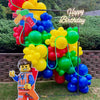 Colorful balloon arch with a 'Happy Birthday' sign and a Lego figure in the foreground