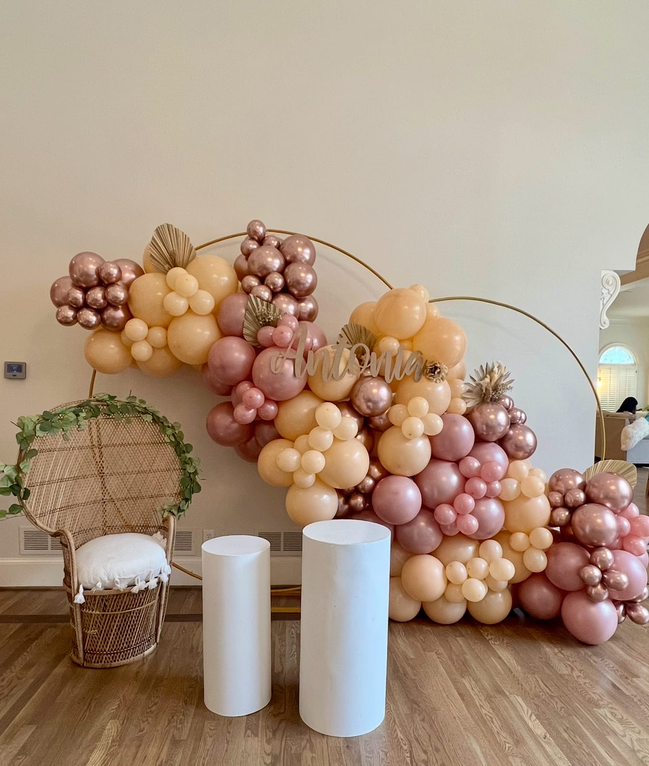 Balloon arch with pink, gold, and white balloons in a room with a wicker chair and white cylindrical tables at a birthday party in Atlanta.