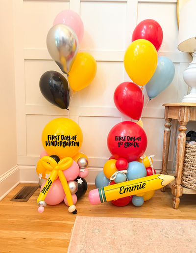 Two colorful balloon clusters styled as graduation and kindergarten birthday decorations on a wooden floor against a white paneled wall. One cluster has yellow, red, pink, black, and silver balloons with 'FIRST DAY OF KINDERGARTEN' text, and the other has red, yellow, light blue, pink, black, and silver balloons with 'FIRST DAY OF GRADUATION' text.