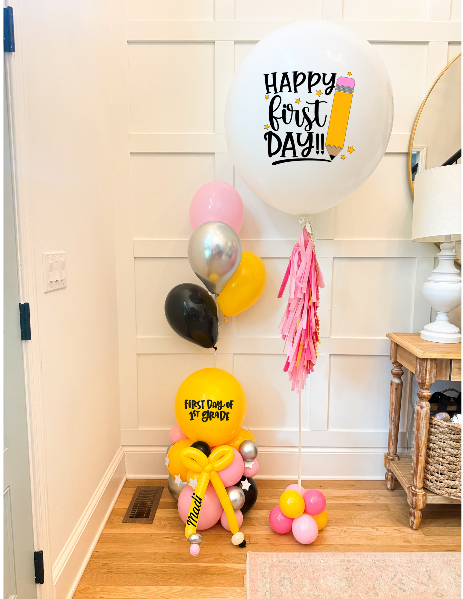 First birthday party balloon cluster in a home hallway, featuring a large white balloon that reads 'Happy First Day!!' with a pencil graphic and pink fringe, a yellow balloon reading 'First Day of School' with a yellow bow, and assorted pink, yellow, black, and metallic silver balloons.