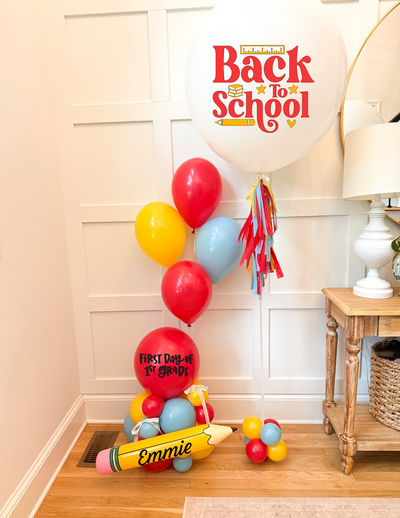 Back to School balloon decoration set in a home interior, featuring a large white balloon that reads 'Back to School' with red lettering, a red balloon that says 'First Day of School,' a yellow novelty pencil, and assorted red, yellow, and light blue balloons.