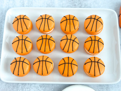 Twelve orange macarons decorated to look like basketballs, arranged in three rows on a white rectangular serving platter.