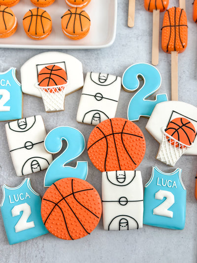 A styled flat-lay of basketball-themed sugar cookies on a light gray surface, including round basketball cookies, number 2 cookies, court-shaped cookies, and blue jersey-shaped cookies with the name Luca and number 2.