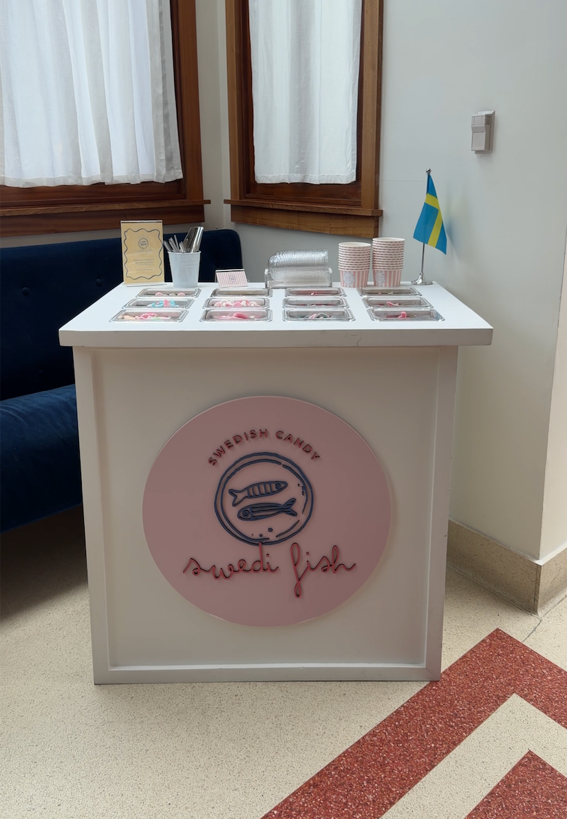 A white candy cart display counter for events around Atlanta with a large pink circular sign reading 'Swedish Candy Sweet Fish' on the front. The counter is topped with multiple small trays of candies, cups, and a small Swedish flag, set in an indoor room with windows, curtains, and a blue chair partially visibilt.
